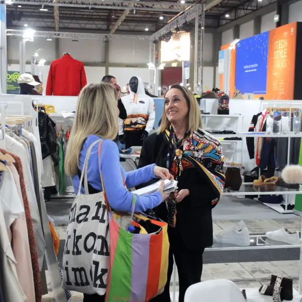 Two people conversing at a clothing trade show with garment displays and mannequins visible in the exhibition hall.