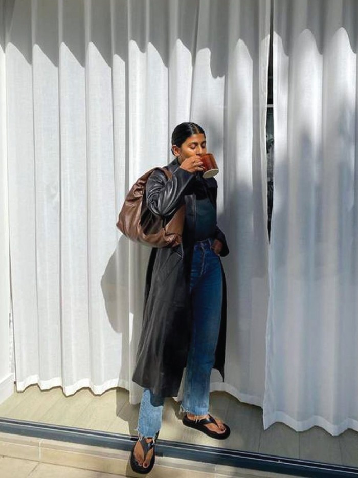 Trends to wear with jeans: long trench coats. Monikh wearing a black long coat with jeans and high-end flip flops.