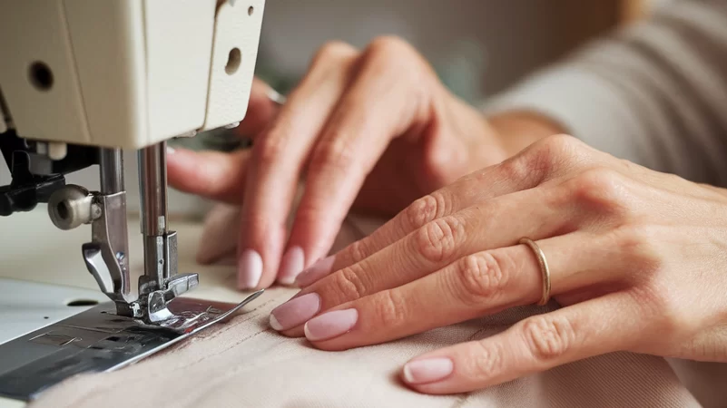 A close-up shows hands guiding light brown fabric under the needle of a white sewing machine.